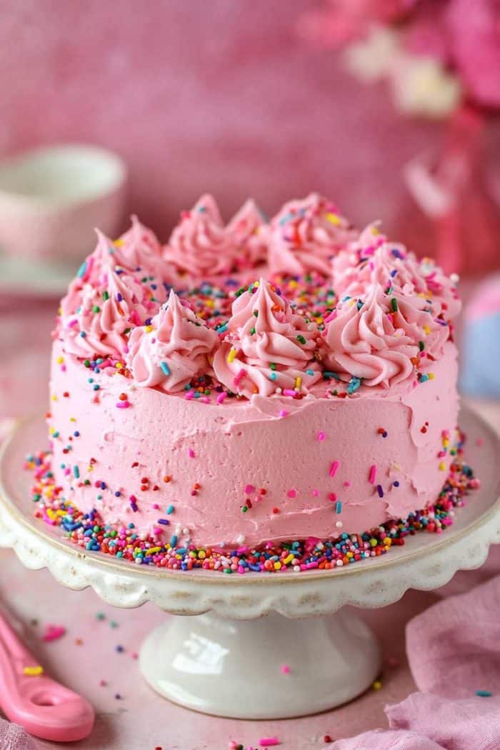 A vibrant pink confetti cake featuring pink sprinkles mixed into the batter before baking. Once cooled, the cake is frosted with light pink buttercream and generously topped with more pink confetti sprinkles on top and around the sides. This playful and festive cake is perfect for birthdays, baby showers, or any joyous occasion.