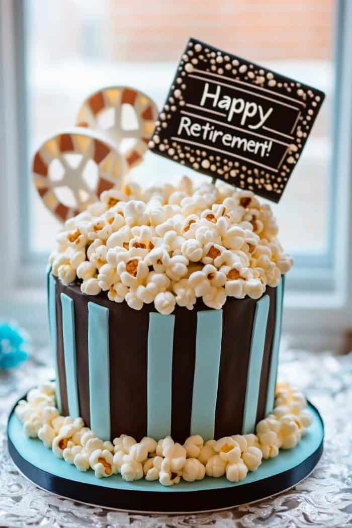 A movie night-themed cake celebrating a retiree's love for cinema, featuring a base of rich chocolate cake layered with caramel buttercream to evoke classic movie theater snacks. The top is decorated with a fondant popcorn bucket overflowing with popcorn made from mini marshmallows lightly dusted with yellow food coloring. Fondant film reels and a movie clapperboard add to the theme, with a personalized message reading 'Happy Retirement!' for a festive touch.