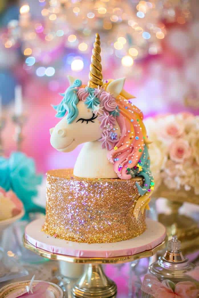 A unicorn-themed cake with a glittery gold base and colorful, intricately designed unicorn head topper, set on a decorated table.