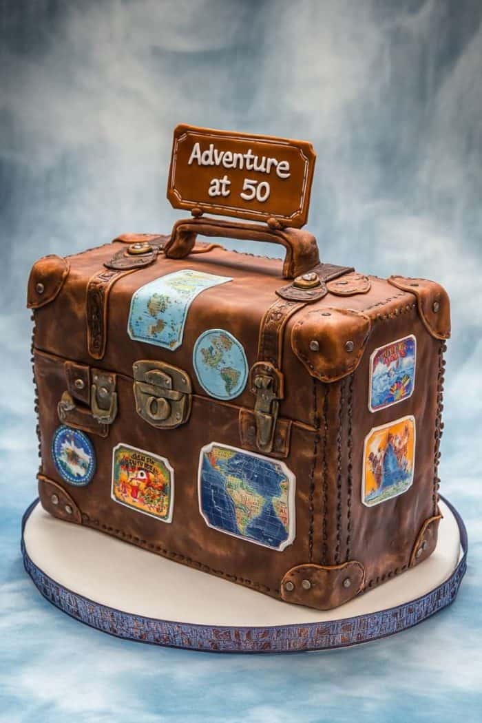 A cake designed as a vintage suitcase decorated with travel-themed stickers. On top, a sign reads "Adventure at 50." The background is a soft blue gradient.