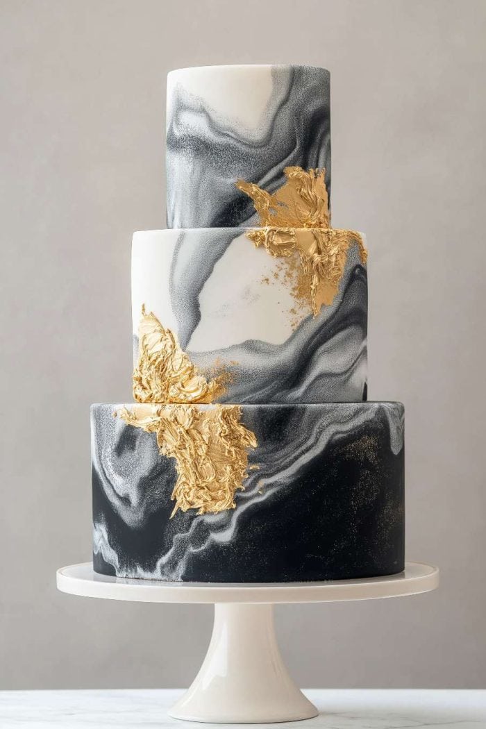 Three-tiered cake with a marble pattern in black, white, and gray, adorned with gold leaf accents. The cake sits on a white stand against a neutral background.