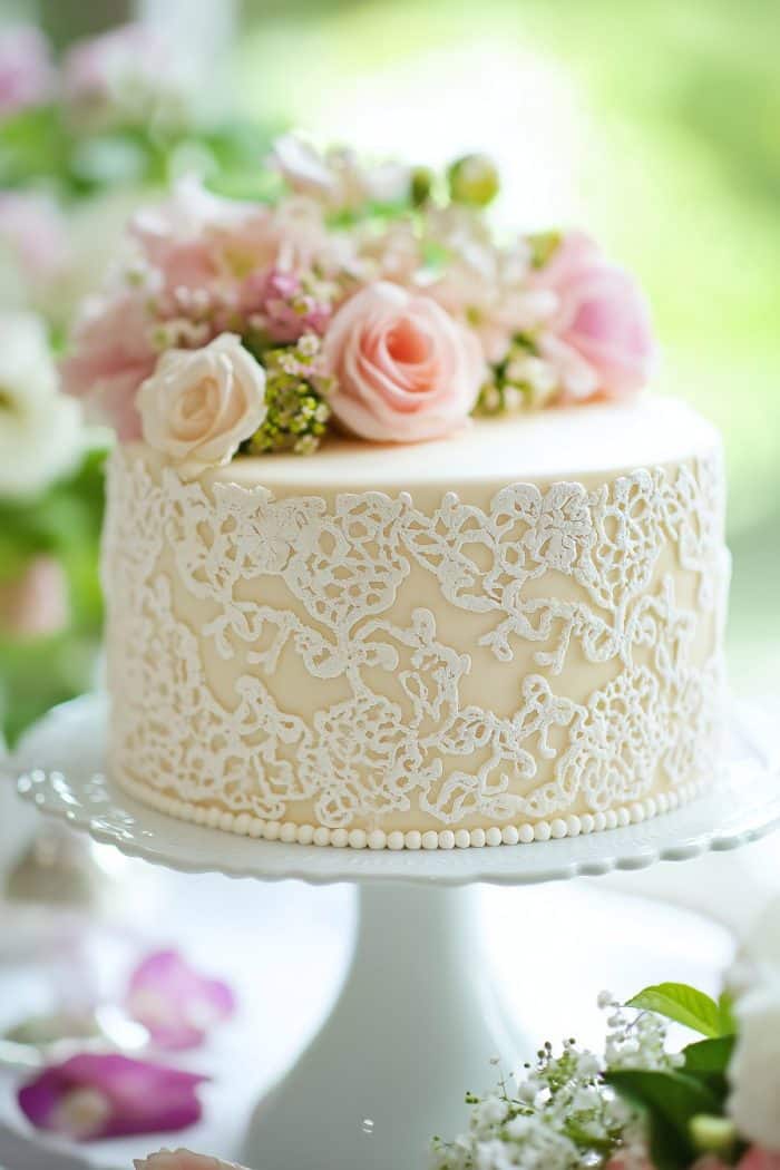 A beautifully decorated cake with intricate white lace icing, topped with pastel pink roses and flowers, sits on a white cake stand. The background is softly blurred with green and pink hues.