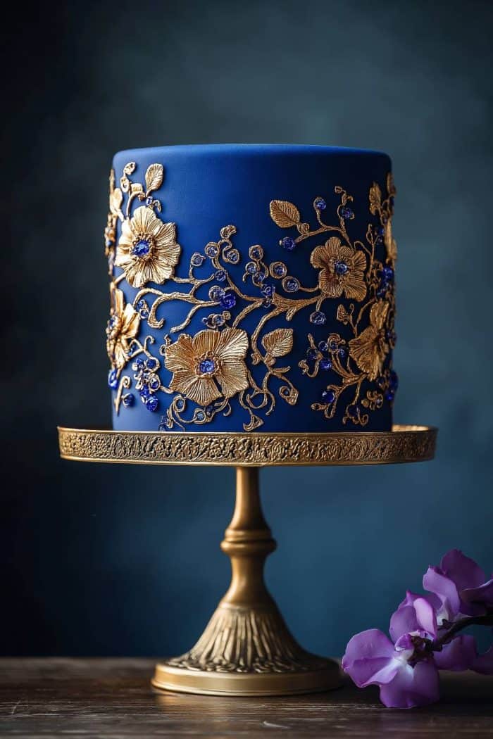 A luxurious blue cake with intricate gold floral designs sits on an ornate gold pedestal. A purple flower lies on the wooden surface next to the cake, creating an elegant and sophisticated presentation.