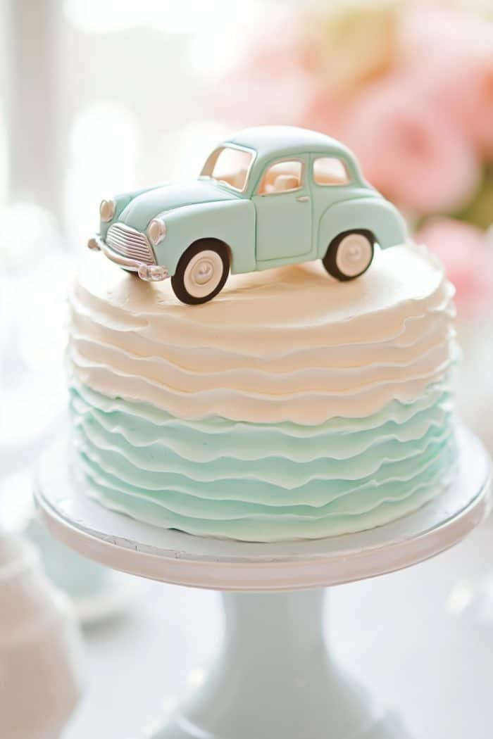 A pastel-themed cake with ombre blue and white icing, topped with a small model of a blue vintage car. The cake is displayed on a white stand.