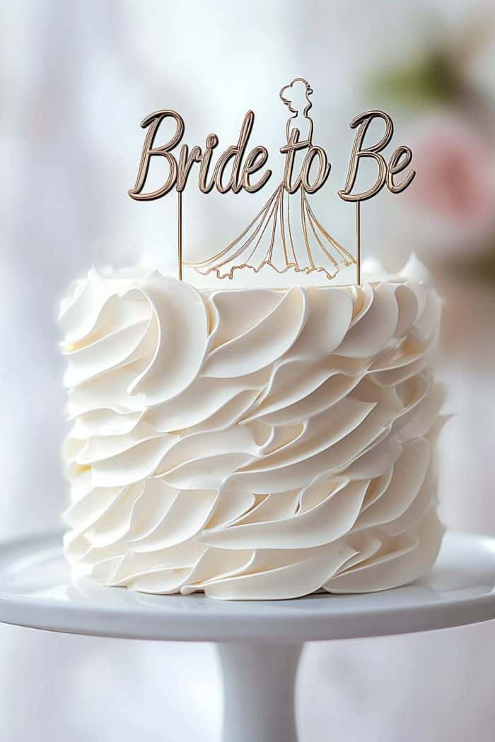 A white cake with elegant wavy frosting, topped with a delicate gold "Bride to Be" decoration featuring a dress silhouette, displayed on a white cake stand.