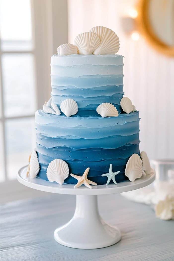 A two-tiered ocean-themed cake with shades of blue, decorated with white seashells and starfish, sits on a white cake stand. The background is softly lit, enhancing the seaside vibe.