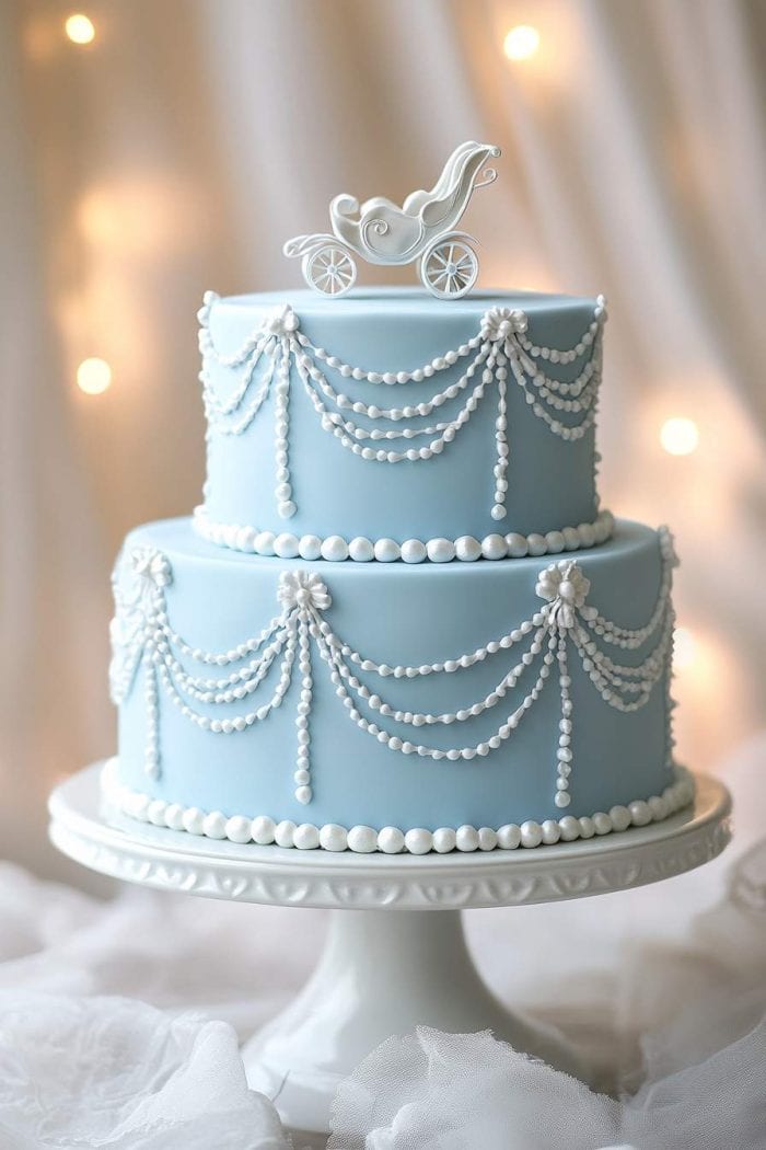 A two-tiered blue cake decorated with white pearl garlands. A white carriage ornament sits on top. The cake is displayed on a white stand with soft, warm lights in the background.