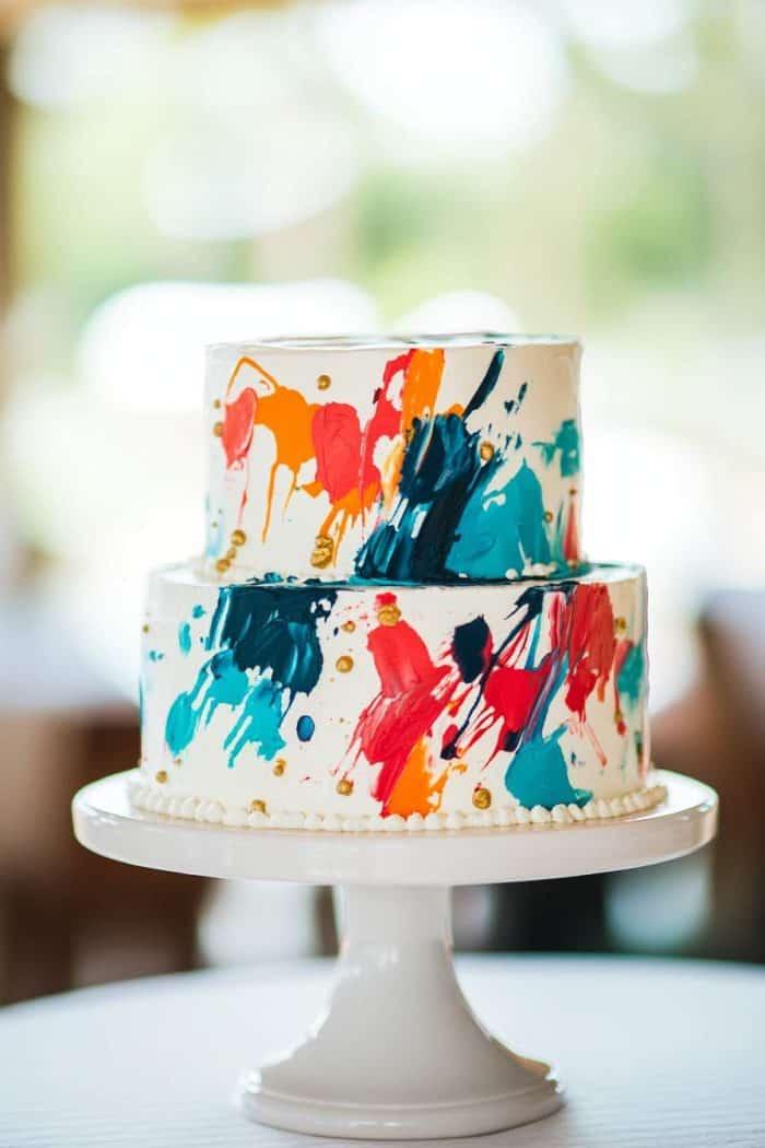 Two-tiered cake on a white stand decorated with colorful brushstroke patterns in red, blue, orange, and teal, accented with small gold details. The background is softly blurred.