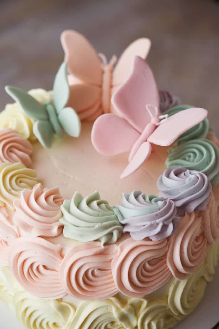 A pastel-colored cake with intricate frosting swirls in shades of pink, purple, and green. Three large, delicate fondant butterflies in pastel pink and mint green are perched on the cake, creating an elegant and whimsical design.