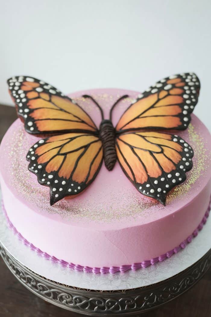 A round cake with light pink frosting is adorned with a large, detailed edible butterfly decoration on top. The butterfly features orange and black wings with white spots. The cake is bordered with purple piping and sits on a decorative stand.