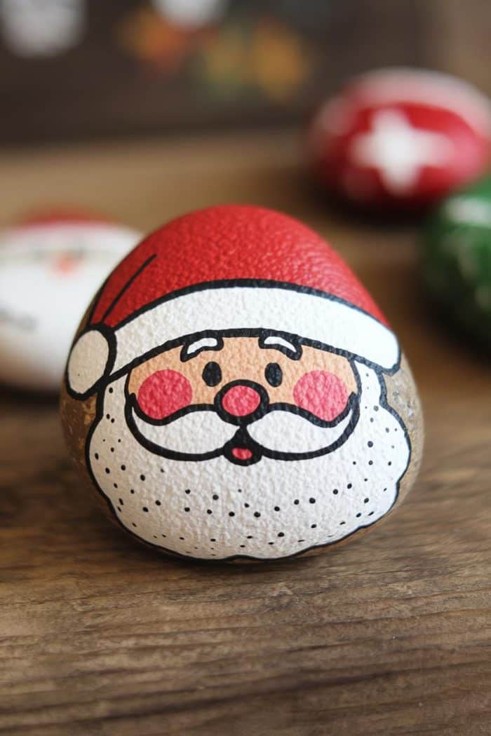 A smooth rock painted with a cheerful Santa Claus face, featuring a red hat and white beard, rests on a wooden surface. Other painted rocks in festive designs are slightly blurred in the background.