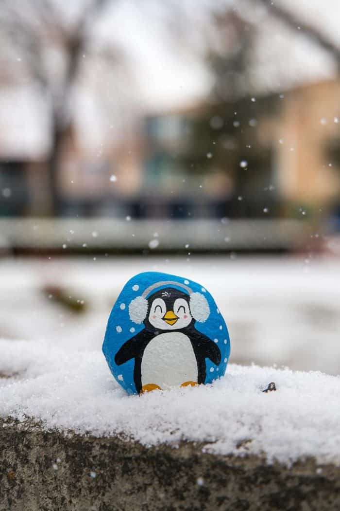 A painted rock featuring a cheerful penguin wearing earmuffs, set against a snowy outdoor background with snowflakes falling. The penguin has a blue backdrop with white dots, enhancing the wintry theme.