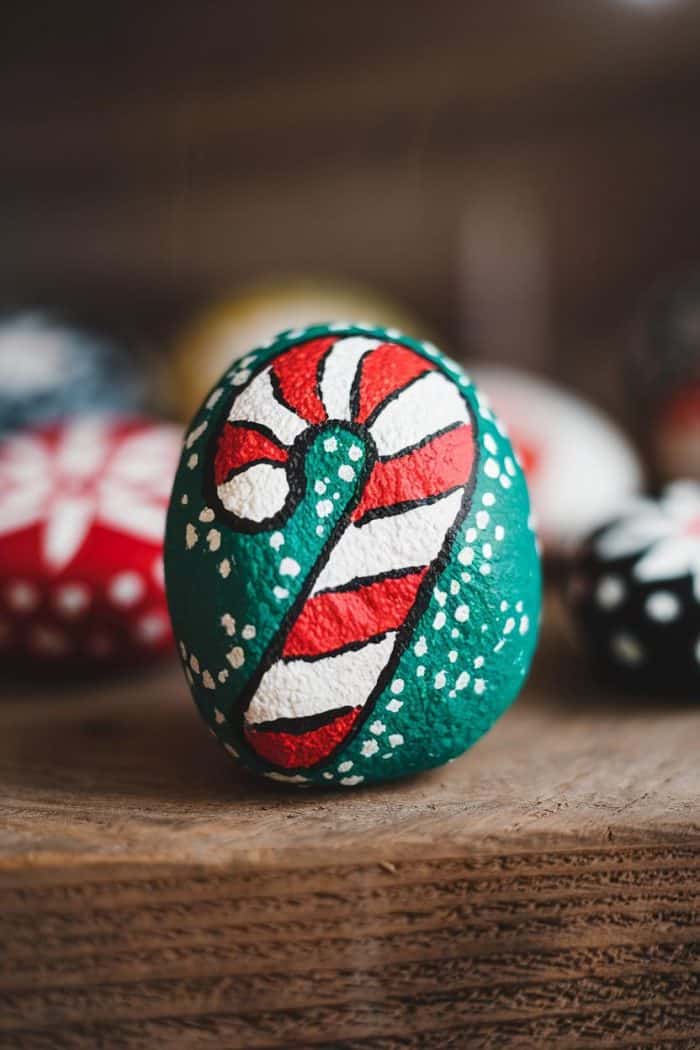 A stone painted with a red and white candy cane design and small white dots on a green background. The stone rests on a wooden surface, surrounded by other blurred, colorfully painted stones.