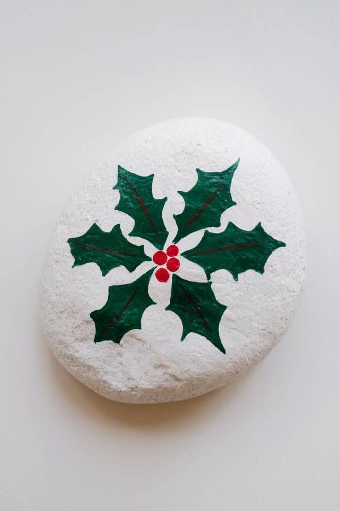 A smooth white stone featuring a painted design of green holly leaves and red berries, symbolizing a festive or holiday theme. The stone is placed on a light, neutral background.