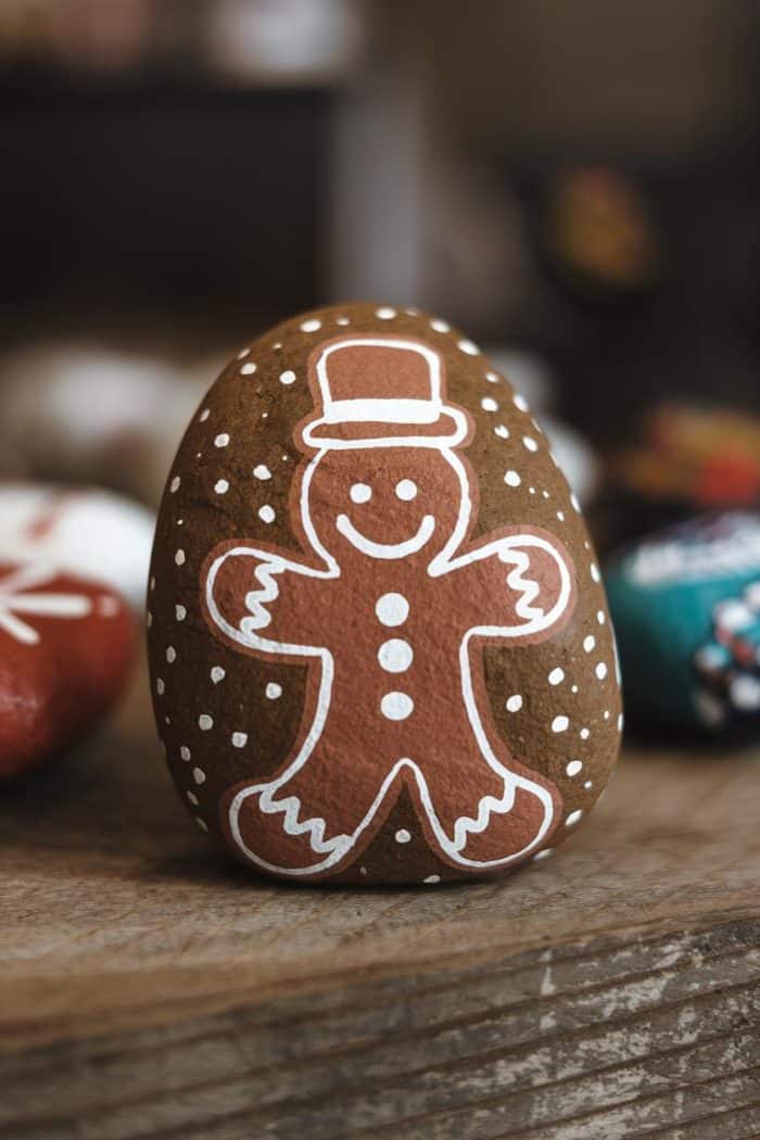A rock painted with a gingerbread man design, featuring a smiling face, top hat, and buttons, is placed on a wooden surface. Other painted rocks can be seen blurred in the background.