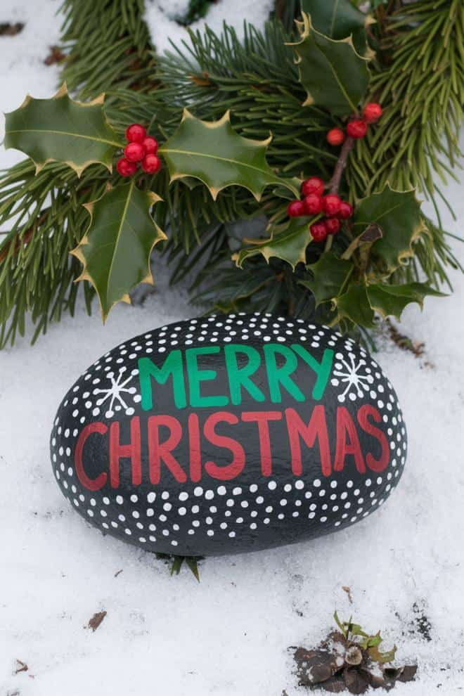 A painted rock on snow with "Merry Christmas" in red and green letters, surrounded by white dots and snowflakes. Pine branches with red berries encircle the rock.