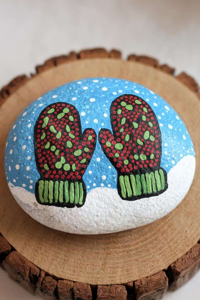 A hand-painted rock displaying two red and green mittens on a blue background with white snowflakes, resting on a wooden slice.