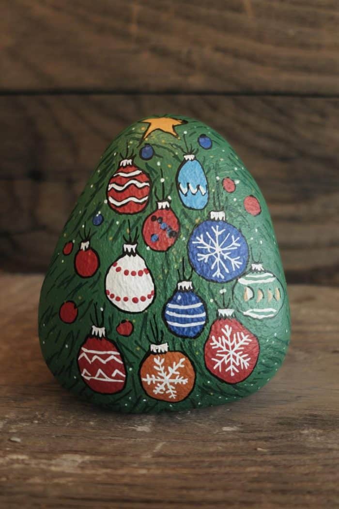 A rock painted to resemble a Christmas tree with colorful ornaments, including snowflakes and geometric patterns, set against a wooden background.
