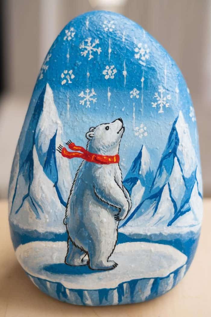 A painted rock featuring a polar bear wearing a red scarf, standing on ice. The background shows snowy mountains and falling snowflakes against a blue sky.