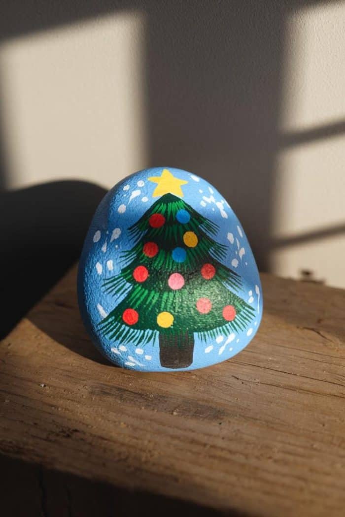 A rock painted with a Christmas tree, adorned with colorful round ornaments and a yellow star on top, sits on a wooden surface. The background is softly lit, creating a warm and festive atmosphere.