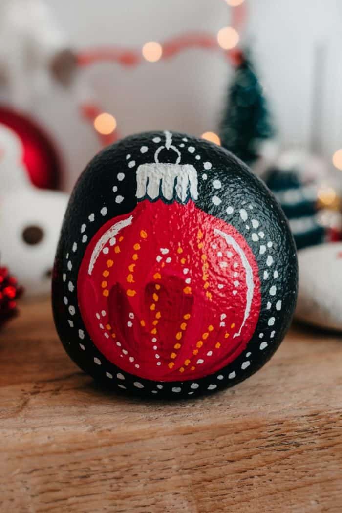 A rock painted with a red Christmas ornament design, featuring white dots and a silver top, rests on a wooden surface. Blurred festive decorations are visible in the background, creating a warm holiday ambiance.