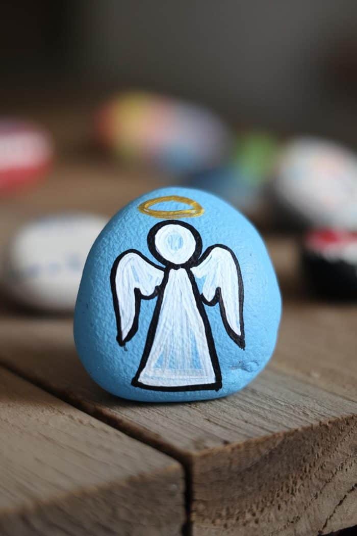 A small, smooth stone painted light blue with a simple white angel design. The angel has a halo and wings. The stone is placed on a wooden surface, with other blurred colorful stones in the background.