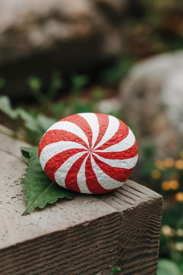 A stone painted with red and white swirls resembling a peppermint sits on a wooden surface, with greenery in the background.