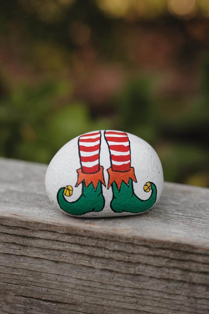 A stone painted with a whimsical design of elf legs with red and white striped socks and green, curled shoes resting on a wooden surface. The background is softly blurred, highlighting the painted detail.