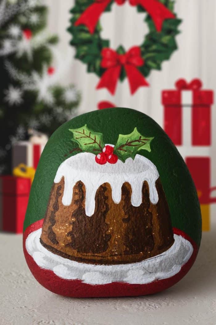 A painted rock features a festive design of a Christmas pudding with white icing and holly on top. In the background, a decorated Christmas tree and wreath are visible, along with stacked gift boxes.