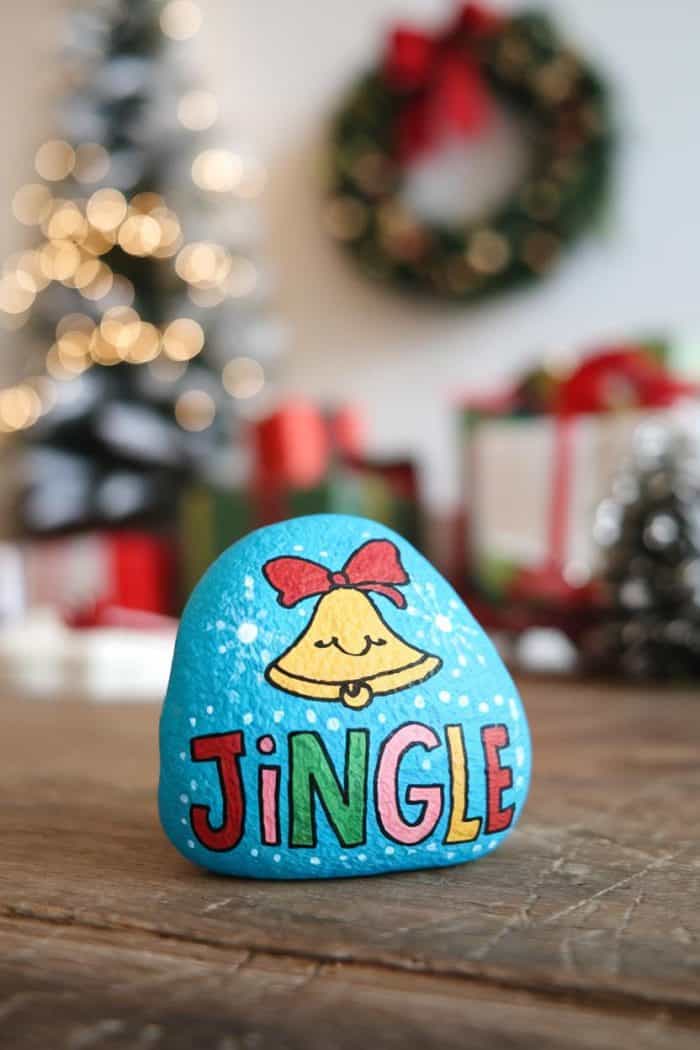 A painted rock with a yellow bell and the word "Jingle" sits on a table. In the background, there is a decorated Christmas tree, a wreath, and blurred holiday lights and gifts.