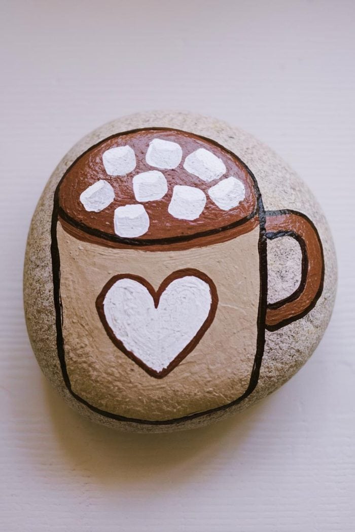 A painted rock with an illustration of a mug filled with hot cocoa and marshmallows. The mug has a heart design on it, and the artwork is on a smooth, round stone.