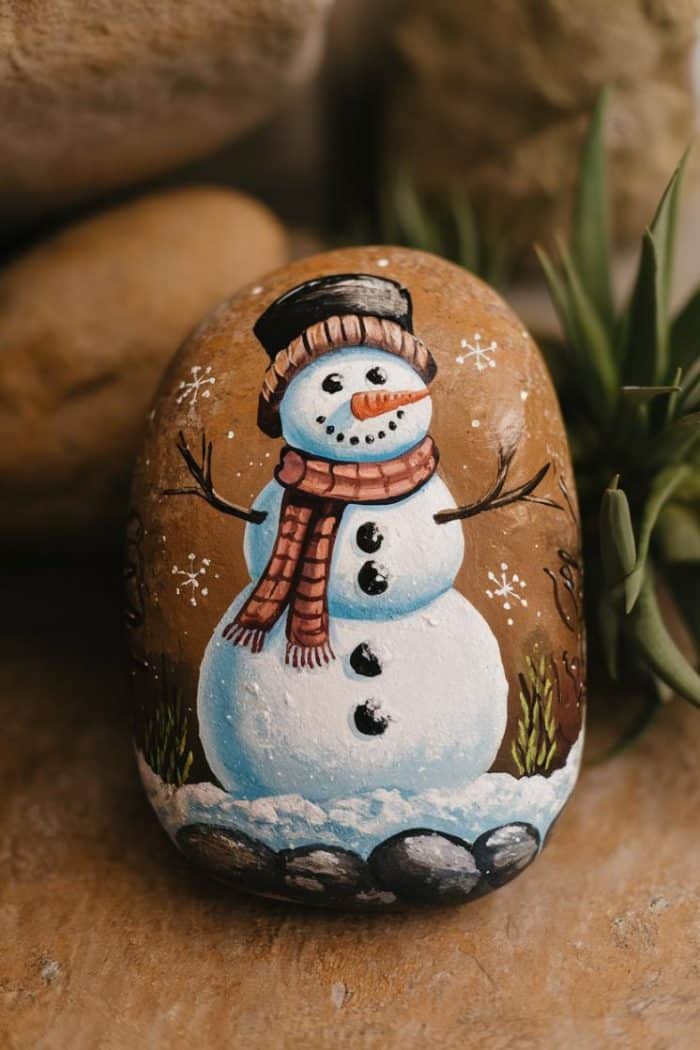 A painted rock depicts a cheerful snowman with a black top hat, red scarf, and carrot nose. It's surrounded by small tufts of grass and snowflakes, sitting on a stone background.