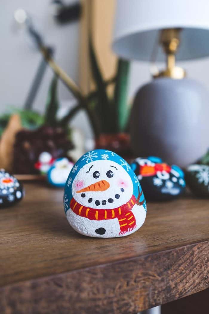 A painted rock featuring a cheerful snowman with a red scarf and carrot nose sits on a wooden surface. Other decorated rocks and a lamp are in the background.