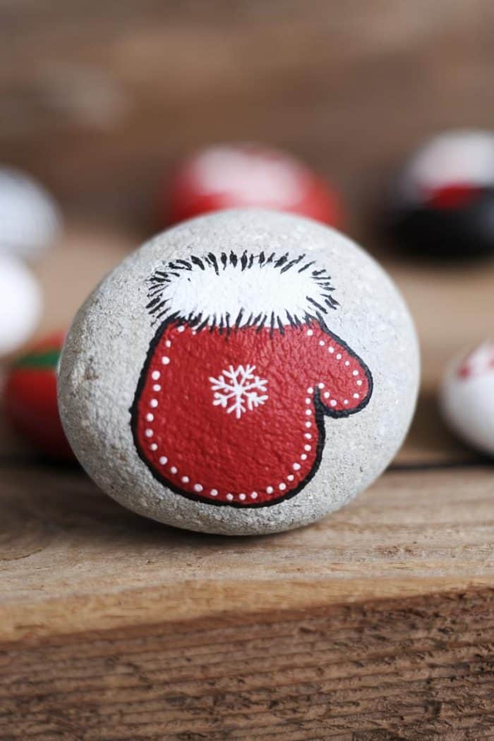 A smooth stone painted with a red mitten featuring a white snowflake design and white trim. The background has blurred, similarly decorated stones.