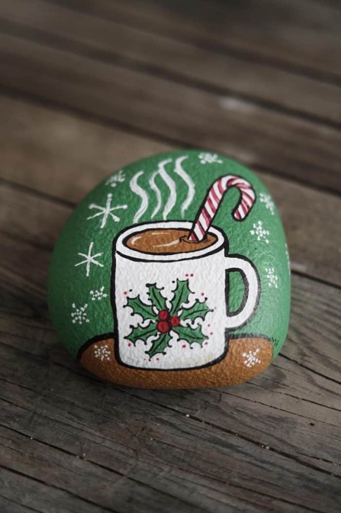 A rock painted with a green background featuring a steaming cup of hot chocolate adorned with a holly design. A candy cane is inside the mug. Snowflakes are scattered around. The rock is placed on a wooden surface.