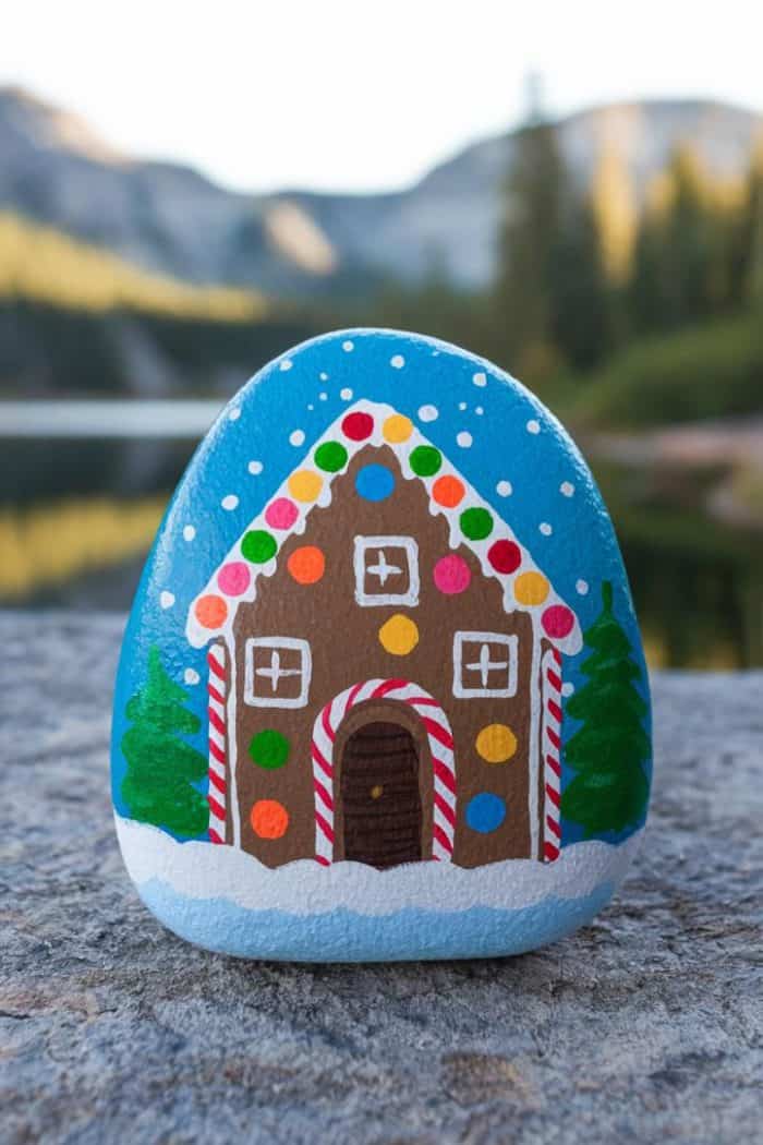 A painted rock with a gingerbread house design featuring a brown house, colorful gumdrops, candy cane borders, snow on the roof, and pine trees. The background shows a blurred landscape with mountains and a lake.
