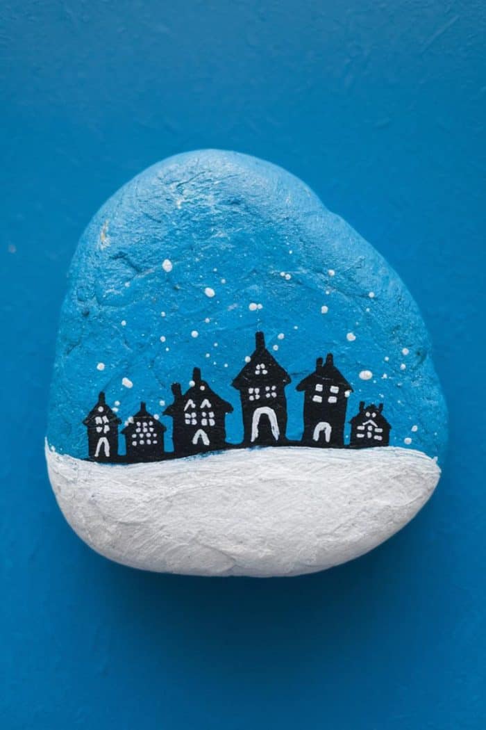 A painted rock with a blue background featuring a snow-covered scene. Five silhouette houses stand against the blue sky, with white snowflakes. The bottom half of the rock is painted white, resembling snow.