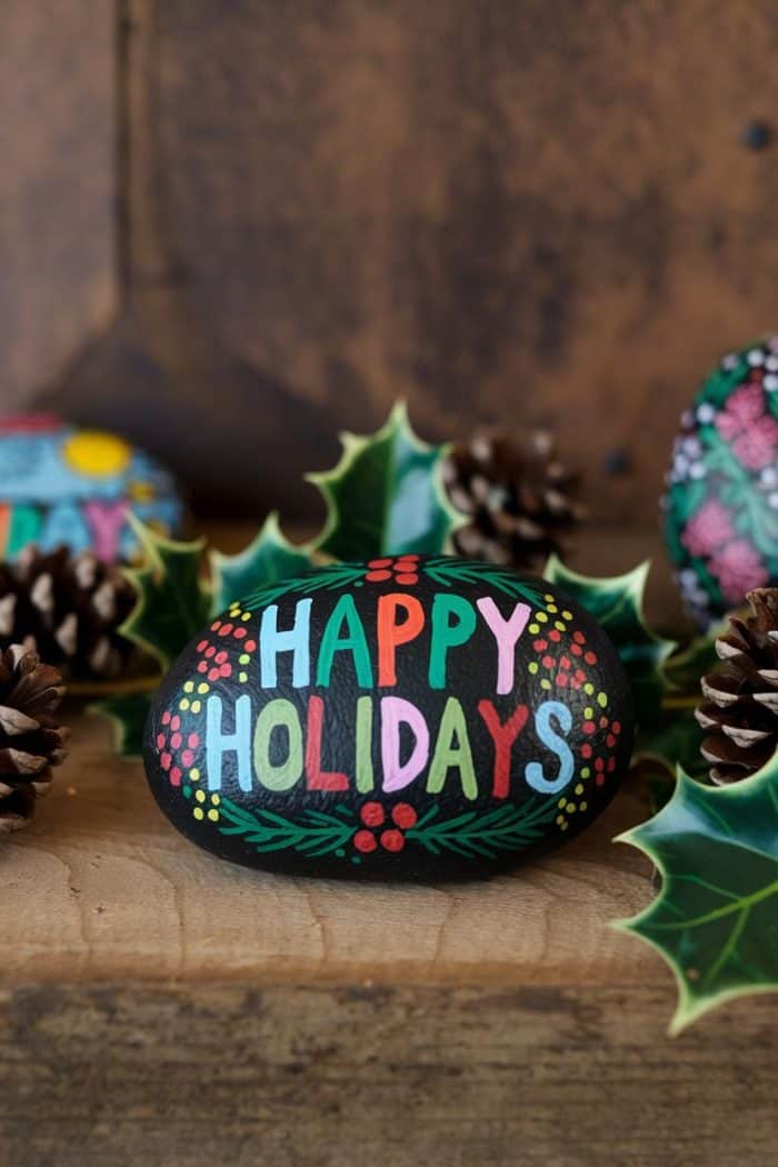 A painted rock with "Happy Holidays" in colorful letters surrounded by pinecones and holly leaves on a wooden surface. Additional decorated rocks are partially visible in the background.