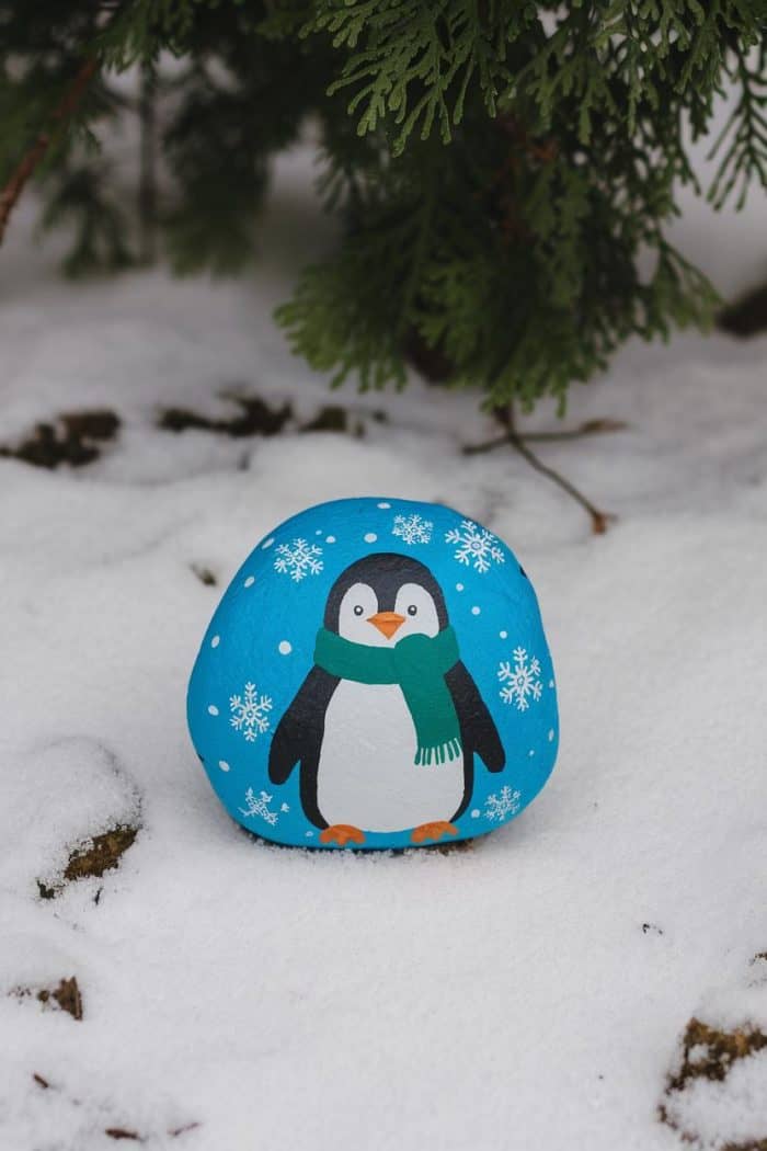 A round stone painted with a cartoon penguin wearing a green scarf, set on snow. Green pine branches are visible in the background.