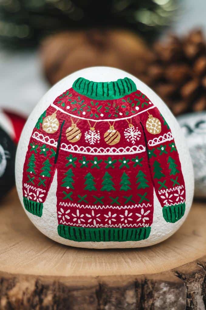 A painted rock featuring a festive red and green Christmas sweater design. The sweater is adorned with patterns of snowflakes, Christmas trees, and ornaments. The rock is placed on a wooden surface with blurred pinecones in the background.