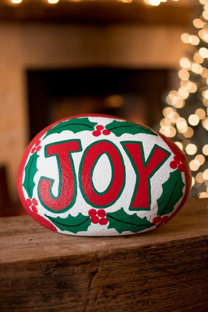 A painted stone with the word "JOY" in bold red letters, surrounded by green holly leaves and red berries. Warm, blurred holiday lights twinkle in the background, creating a festive atmosphere.
