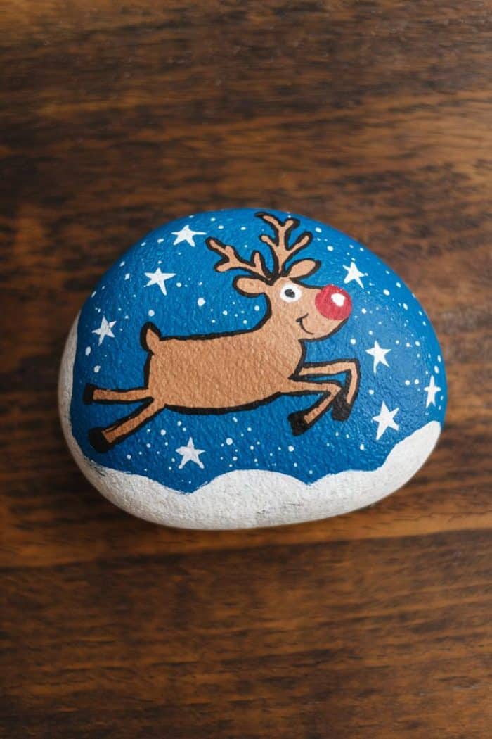 A painted rock featuring a reindeer with a red nose and antlers, leaping against a blue background with white stars. The lower part of the rock is painted white, resembling snow.