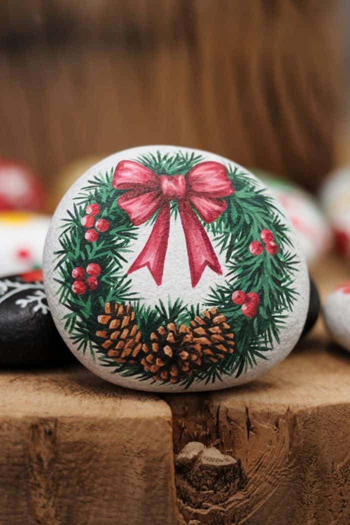 A stone painted with a Christmas wreath featuring a red bow, pinecones, and holly berries, resting on a wooden surface.