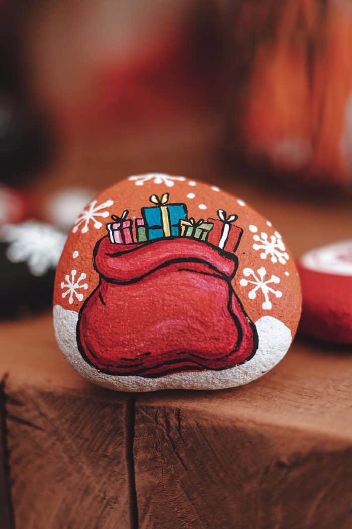A painted rock with a red sack filled with colorful gift boxes, surrounded by white snowflakes, rests on a wooden surface. The background is softly blurred, enhancing the festive theme.