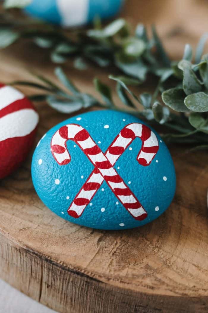 A blue painted rock with white dots features two crossed red and white candy canes. The rock rests on a wooden surface, surrounded by green foliage in the background.