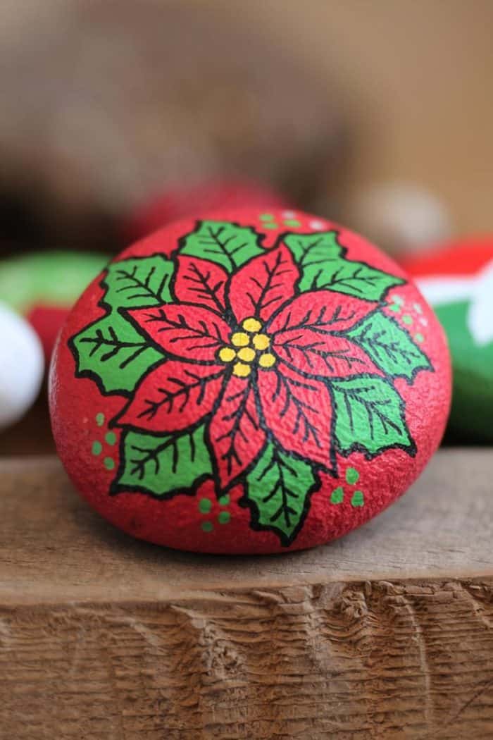 A round stone painted with a vibrant red poinsettia design, accented by green leaves and yellow dots in the center, is displayed on a wooden surface.