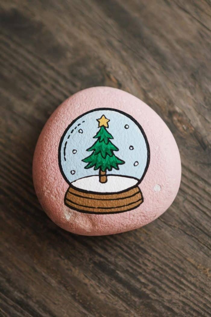 A painted rock featuring a snow globe with a decorated Christmas tree inside. The rock is placed on a rustic wooden surface.