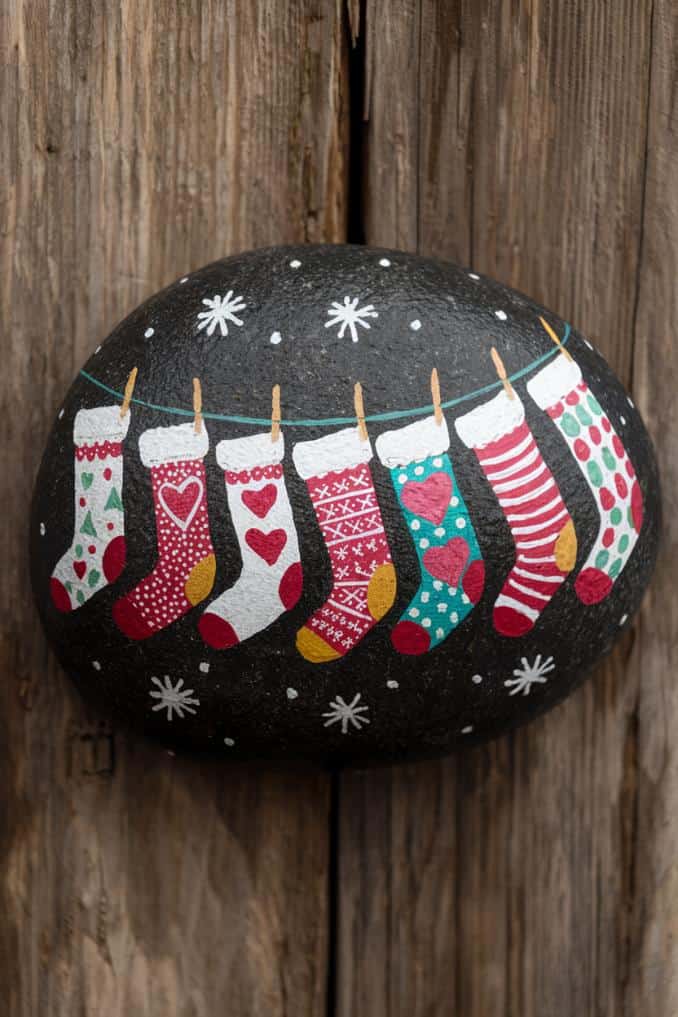 A painted rock displays colorful Christmas stockings hanging on a line against a dark background, with snowflakes scattered around. Each stocking has a unique pattern, including hearts, polka dots, and stripes. The rock rests on a wooden surface.