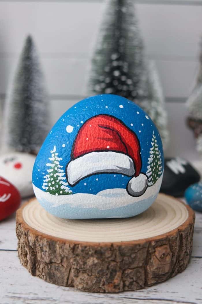 A painted rock depicting a Santa hat on a snowy background with pine trees, resting on a wooden slice. Several snowy decorative trees are blurred in the background.