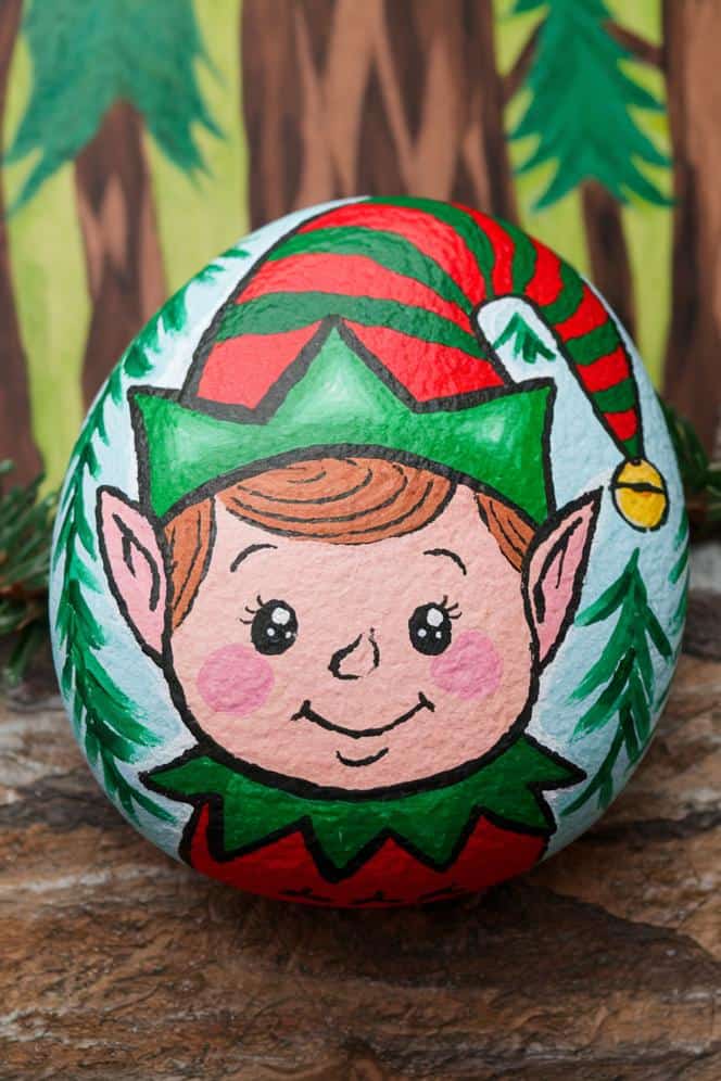 A painted rock features a smiling elf with rosy cheeks and pointed ears, wearing a green and red hat. The background includes green trees on the rock and a wooden backdrop.
